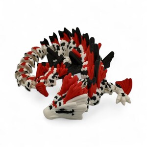 May include: A detailed, three-dimensional dragon figurine. The dragon features a white head with black spots and red accents. The body is a mix of red, black, and white, with a curved tail. The figurine is set against a plain white background.