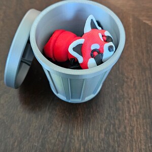 May include: A 3D-printed red panda figurine inside a small, gray trash can with the lid slightly ajar. The red panda has white and black markings. The trash can is on a dark brown wooden surface.
