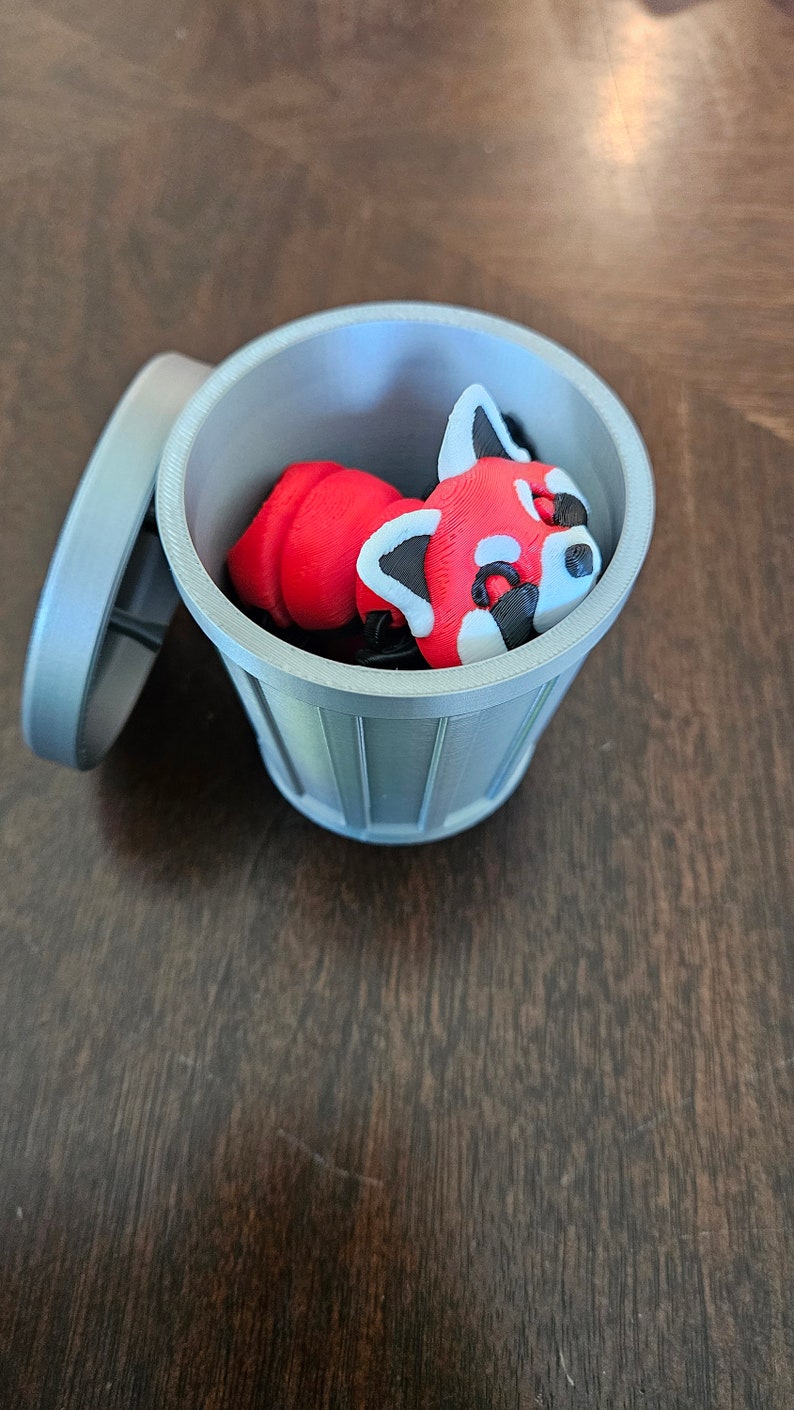 May include: A 3D-printed red panda figurine inside a small, gray trash can with the lid slightly ajar. The red panda has white and black markings. The trash can is on a dark brown wooden surface.