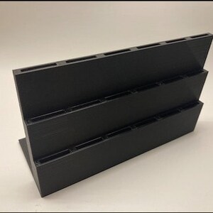 May include: Black plastic organizer with three tiers and multiple slots. The organizer is designed to hold small items, such as pens, pencils, or paintbrushes.