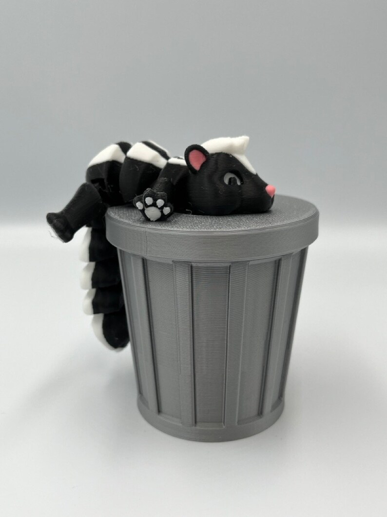 May include: A 3D-printed skunk figurine with black and white markings, resting on a gray trash can. The skunk has a pink nose and ears. The trash can is cylindrical with vertical lines.
