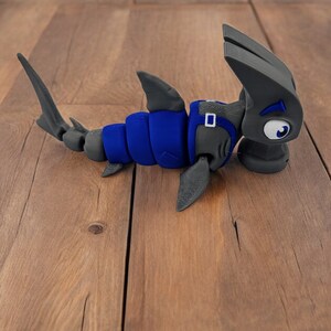 May include: A blue and gray 3D printed hammerhead shark toy. The shark has a large head with a hammer-shaped snout and a blue body with gray fins.