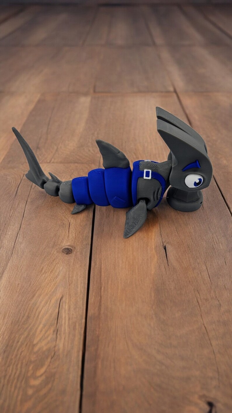 May include: A blue and gray 3D printed hammerhead shark toy. The shark has a large head with a hammer-shaped snout and a blue body with gray fins.