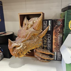 May include: A gold dragon head bookend with a textured surface. The bookend is holding up two board games, "One Night Ultimate Werewolf" and "Daybreak".