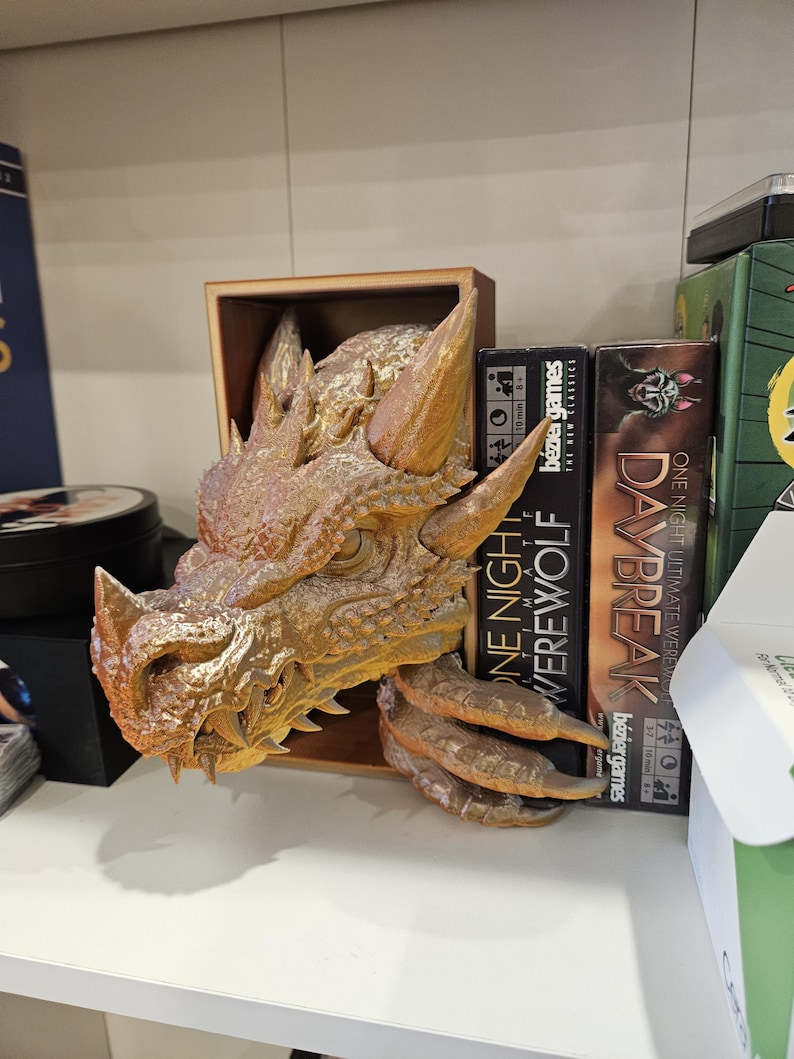 May include: A gold dragon head bookend with a textured surface. The bookend is holding up two board games, "One Night Ultimate Werewolf" and "Daybreak".