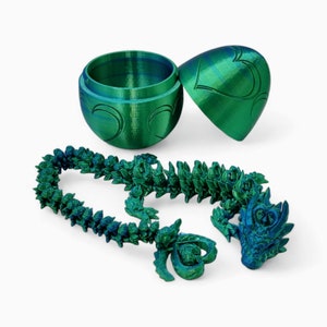 May include: A green and blue iridescent dragon necklace with a matching egg-shaped box. The dragon is made of plastic and has a detailed design. The box is made of plastic and has a heart design.