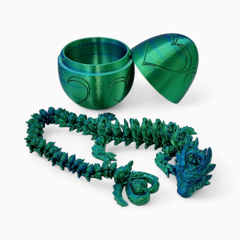 May include: A green and blue iridescent dragon necklace with a matching egg-shaped box. The dragon is made of plastic and has a detailed design. The box is made of plastic and has a heart design.