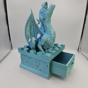 May include: A teal-colored dragon figurine with outstretched wings and an open mouth sits atop a castle-like box with a drawer. The dragon has textured scales and a detailed wing design. The box has a brick-like texture.