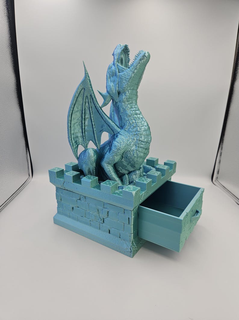 May include: A teal-colored dragon figurine with outstretched wings and an open mouth sits atop a castle-like box with a drawer. The dragon has textured scales and a detailed wing design. The box has a brick-like texture.