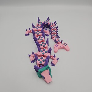 May include: A pink and purple articulated dragon figurine. The dragon's body is made of pink segments with blue button details, connected by purple spines. The head is pink and green, and the tail ends in a pink controller.