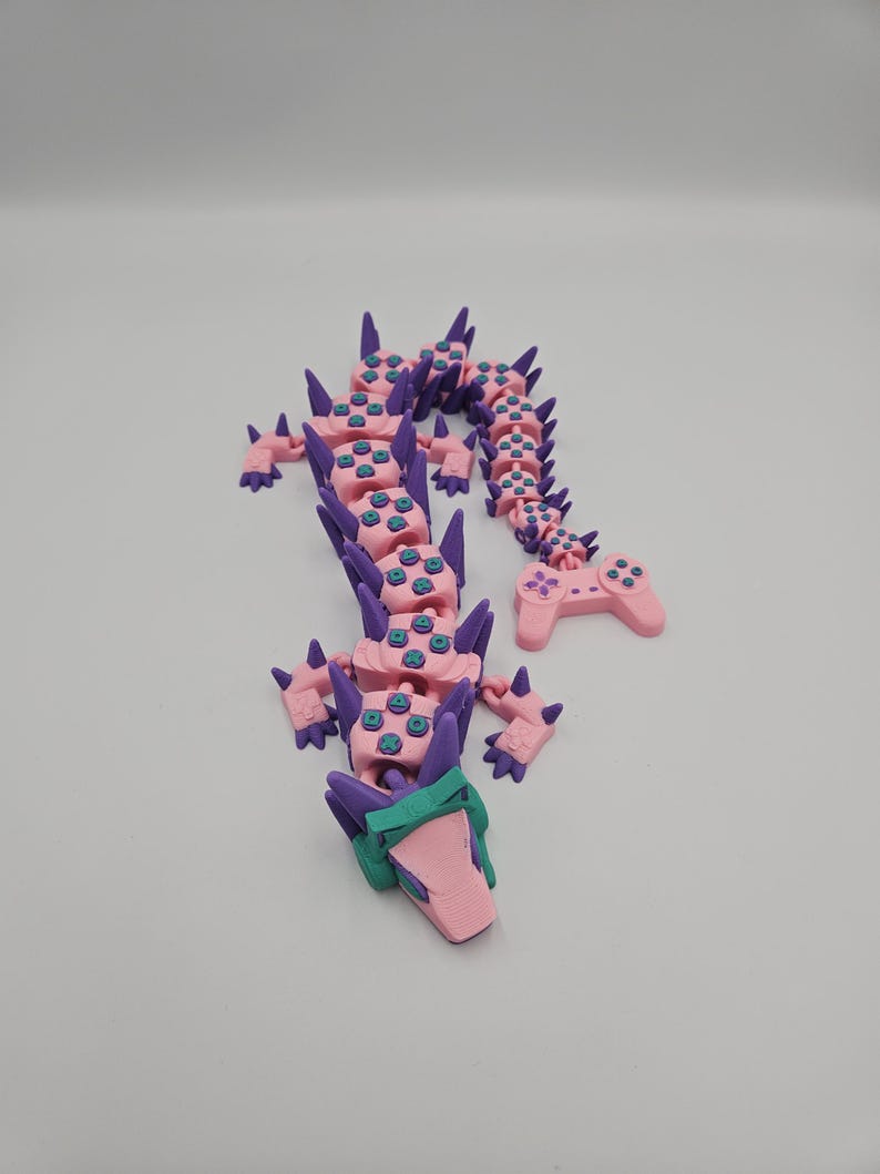 May include: A pink and purple articulated dragon figurine. The dragon's body is made of pink segments with blue button details, connected by purple spines. The head is pink and green, and the tail ends in a pink controller.