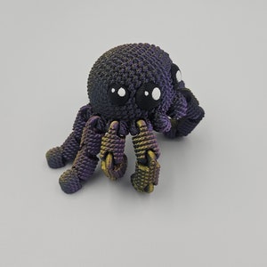 Op de afbeelding: Een kleine, paarse en gouden 3D-geprinte octopus met witte ogen. De octopus heeft acht tentakels en staat op een wit oppervlak.