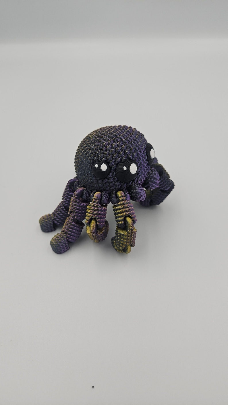 Op de afbeelding: Een kleine, paarse en gouden 3D-geprinte octopus met witte ogen. De octopus heeft acht tentakels en staat op een wit oppervlak.