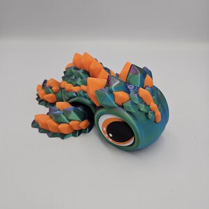 Pode incluir: Figura de drag&atilde;o articulada impressa em 3D. O drag&atilde;o apresenta um corpo azul-petr&oacute;leo e azul com detalhes em laranja e verde. Possui um olho grande com uma pupila preta e um destaque branco. O drag&atilde;o est&aacute; em uma superf&iacute;cie branca.