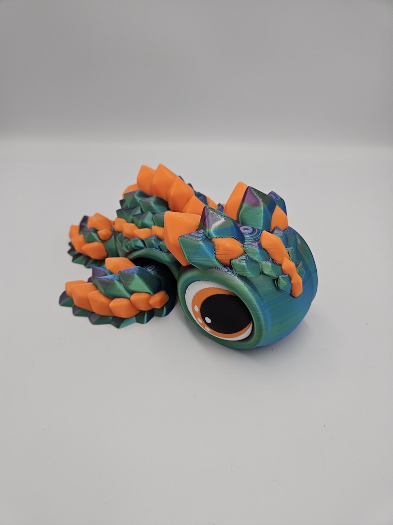 Pode incluir: Figura de drag&atilde;o articulada impressa em 3D. O drag&atilde;o apresenta um corpo azul-petr&oacute;leo e azul com detalhes em laranja e verde. Possui um olho grande com uma pupila preta e um destaque branco. O drag&atilde;o est&aacute; em uma superf&iacute;cie branca.