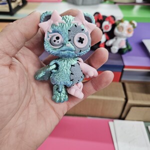 Puede incluir: Peque&ntilde;a figura articulada de oso con est&eacute;tica desgastada. El oso presenta un cuerpo turquesa con parches grises, detalles rosas y ojos de bot&oacute;n. Tiene lazos rosas en las orejas y una cruz cosida en la boca. Otras figuras similares son visibles en el fondo.