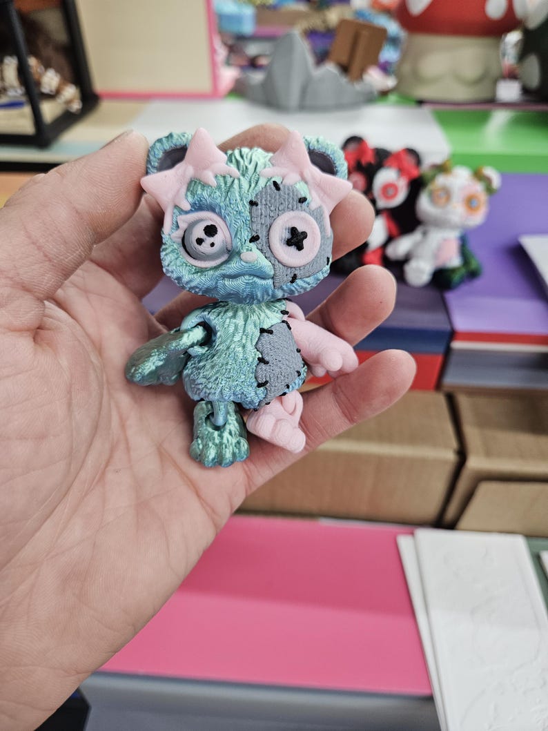 Puede incluir: Peque&ntilde;a figura articulada de oso con est&eacute;tica desgastada. El oso presenta un cuerpo turquesa con parches grises, detalles rosas y ojos de bot&oacute;n. Tiene lazos rosas en las orejas y una cruz cosida en la boca. Otras figuras similares son visibles en el fondo.