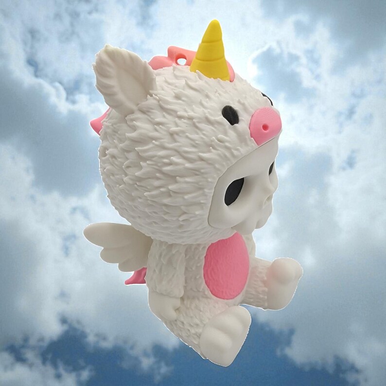 Puede incluir: Figura blanca de una calavera con un disfraz de unicornio con cuerno amarillo, detalles rosas y peque&ntilde;as alas blancas. La figura tiene ojos negros sobre un fondo de cielo azul nublado.