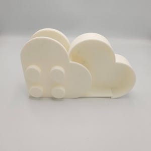 May include: A cream-colored, cloud-shaped napkin holder. The holder has a rounded design with four circular details on the front. The holder is made of a solid material and has a smooth surface. The design is modern and minimalist.