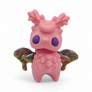 May include: A pink, stylized figurine with large purple eyes and iridescent wings. The figure has a textured surface and a whimsical design, resembling a creature or character. The figurine stands on two legs and has a unique, artistic appearance.