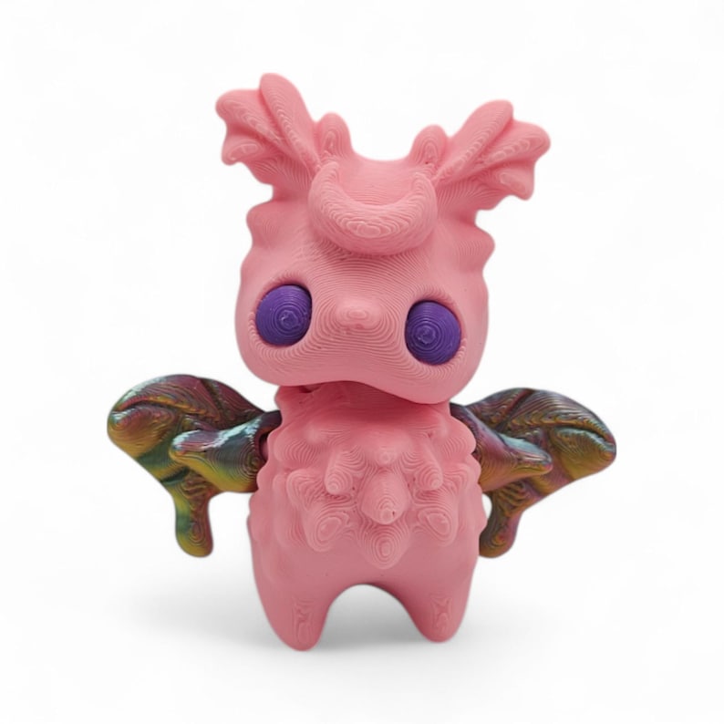 May include: A pink, stylized figurine with large purple eyes and iridescent wings. The figure has a textured surface and a whimsical design, resembling a creature or character. The figurine stands on two legs and has a unique, artistic appearance.