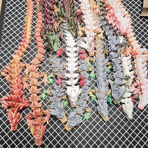 May include: A collection of 10 colourful 3D printed dragon figurines with intricate details. Each dragon is made of a different colour plastic and has a unique design. The dragons are arranged in a row on a black grid background.