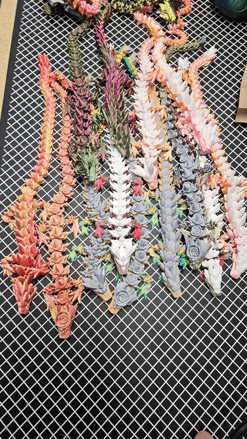 May include: A collection of 10 colourful 3D printed dragon figurines with intricate details. Each dragon is made of a different colour plastic and has a unique design. The dragons are arranged in a row on a black grid background.