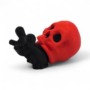 May include: A red skull figurine with black eye sockets and a black hand grasping its jaw. The skull is the focal point, with the hand appearing to hold or interact with it. The design is a unique, artistic representation.