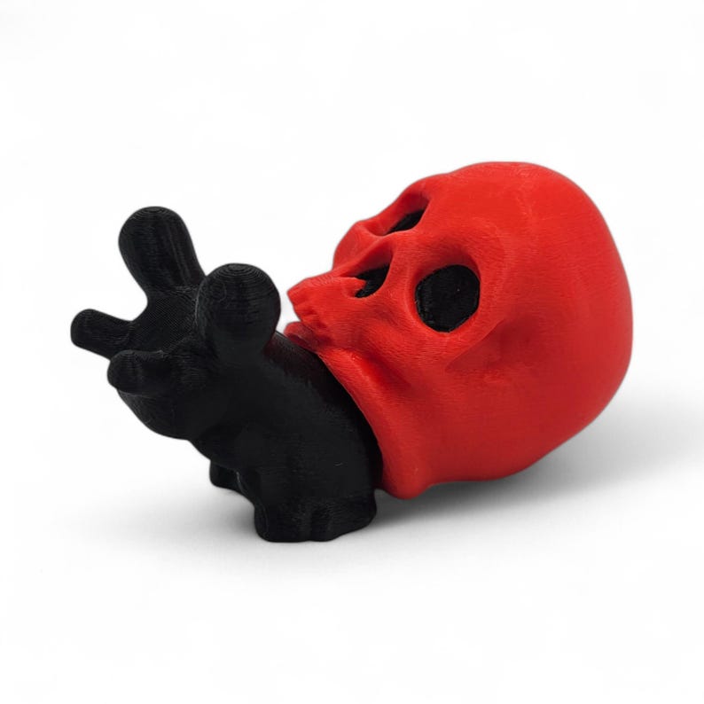 May include: A red skull figurine with black eye sockets and a black hand grasping its jaw. The skull is the focal point, with the hand appearing to hold or interact with it. The design is a unique, artistic representation.