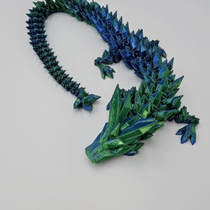 May include: A 3D printed dragon figurine with a green and blue iridescent finish. The dragon is posed in a curled position with its head resting on its front legs. The dragon's scales are detailed and textured.