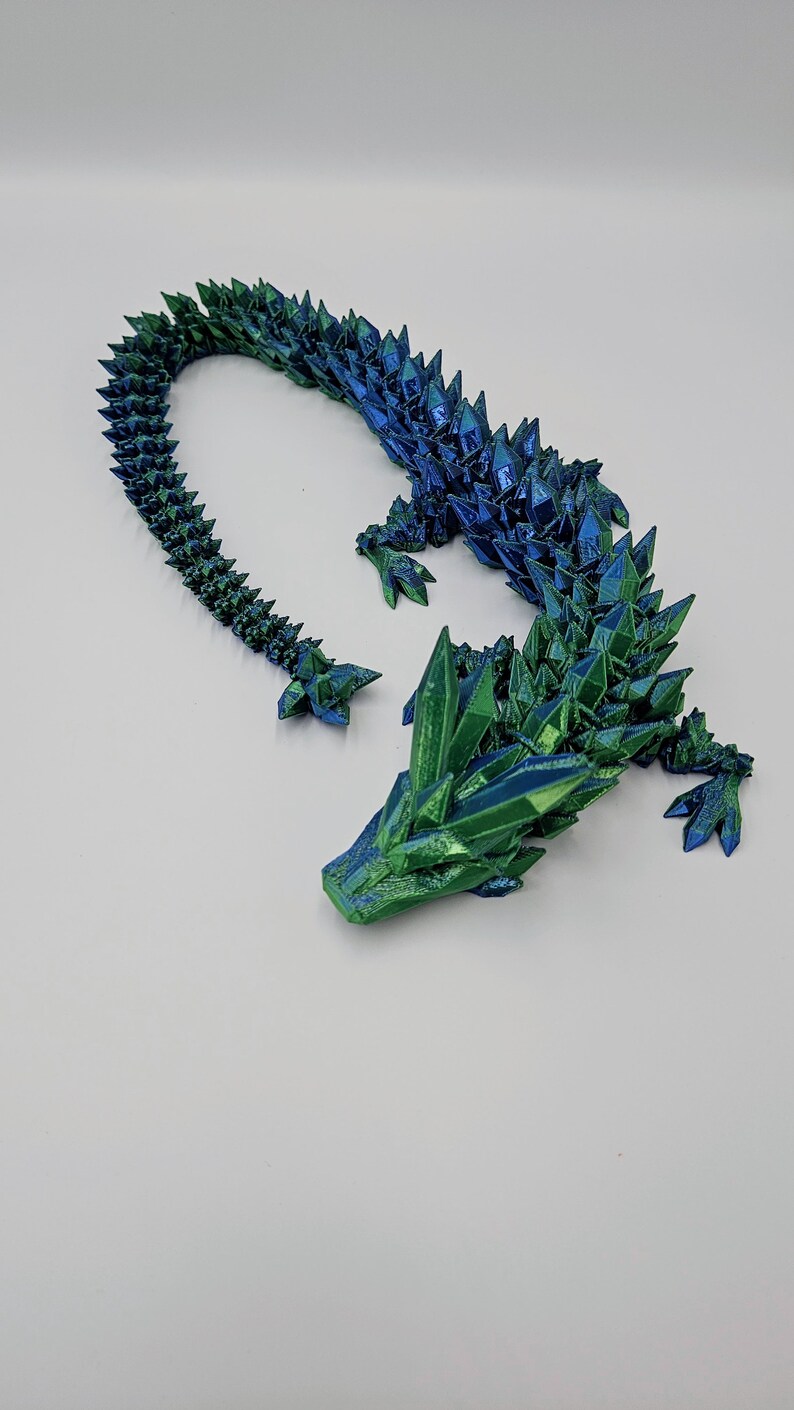 May include: A 3D printed dragon figurine with a green and blue iridescent finish. The dragon is posed in a curled position with its head resting on its front legs. The dragon's scales are detailed and textured.