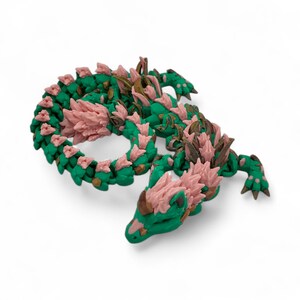 May include: A detailed, 3D-printed dragon figurine. The dragon features a green body with pink and gold accents. The design includes intricate scales and a stylized, curled pose, showcasing the creature's form.