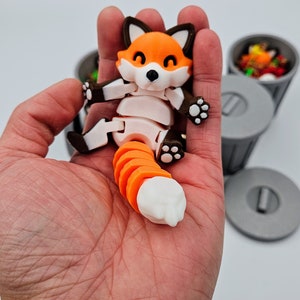 May include: A 3D-printed articulated fox figurine with an orange and white segmented tail. The fox has an orange body, white belly, and brown accents. The fox is held in a hand, with small gray trash cans in the background.