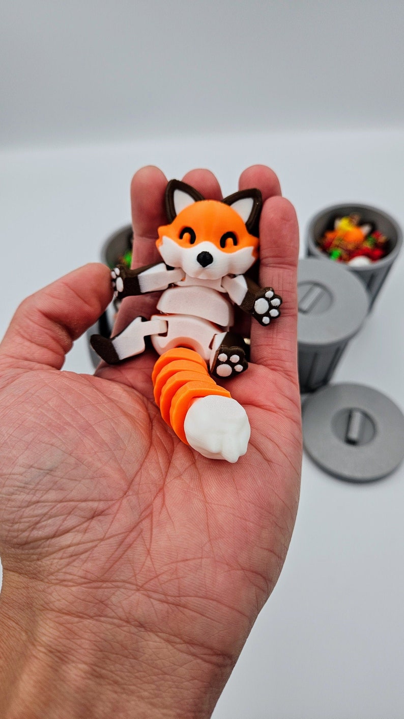 May include: A 3D-printed articulated fox figurine with an orange and white segmented tail. The fox has an orange body, white belly, and brown accents. The fox is held in a hand, with small gray trash cans in the background.
