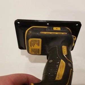May include: A black and yellow cordless drill is mounted to a black plastic wall mount. The drill has a black and yellow handle and a black trigger. The wall mount has four screw holes.