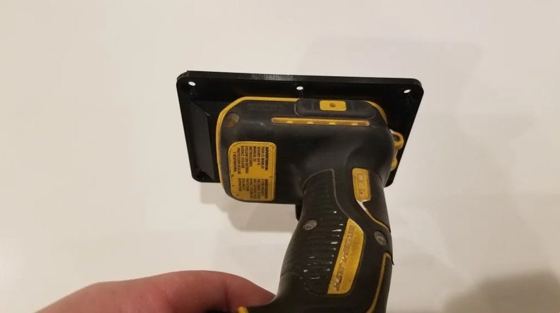 May include: A black and yellow cordless drill is mounted to a black plastic wall mount. The drill has a black and yellow handle and a black trigger. The wall mount has four screw holes.