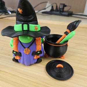 Puede incluir: Una caprichosa figura de gnomo de Halloween con un sombrero de bruja negro con detalles en naranja y una banda verde. El gnomo tiene trenzas naranjas, un vestido morado y est&aacute; junto a un caldero negro con una escoba y una tapa. Decoraci&oacute;n de Halloween.