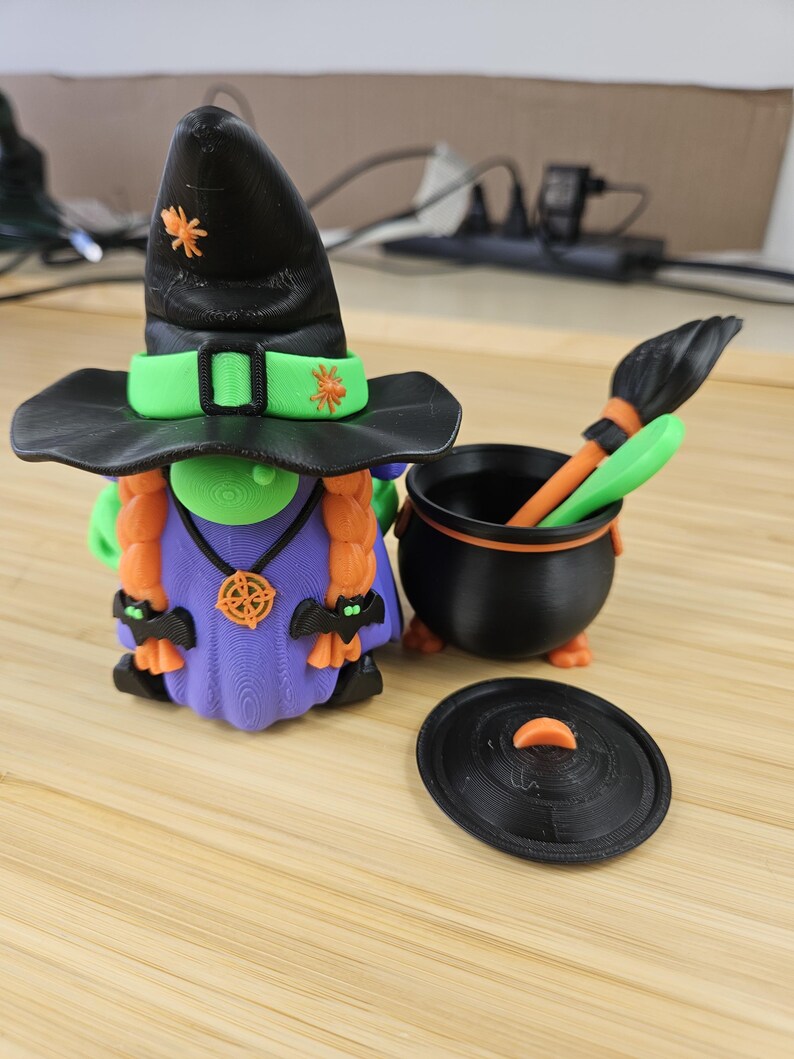 Puede incluir: Una caprichosa figura de gnomo de Halloween con un sombrero de bruja negro con detalles en naranja y una banda verde. El gnomo tiene trenzas naranjas, un vestido morado y est&aacute; junto a un caldero negro con una escoba y una tapa. Decoraci&oacute;n de Halloween.