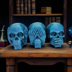 Op de afbeelding: Drie blauwe schedelsculpturen op een houten tafel. Elke schedel vertegenwoordigt "Zie geen kwaad, hoor geen kwaad, spreek geen kwaad." De schedels hebben gedetailleerde kenmerken en zijn een uniek decoratief item.