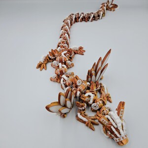 May include: A 3D printed, articulated dragon sculpture with a copper and white colour scheme. The dragon is posed in a crawling position with its head raised.