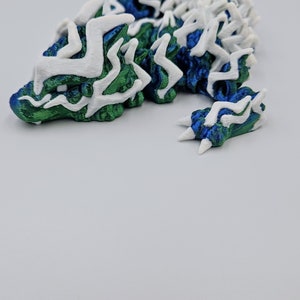May include: A 3D printed dragon figurine with a white and green patterned design. The dragon is curled up in a resting position with its head resting on its front paws.