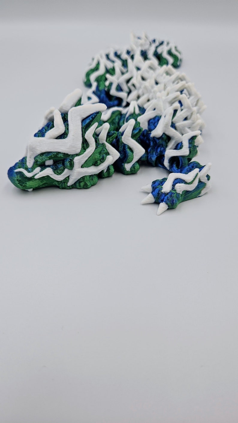 May include: A 3D printed dragon figurine with a white and green patterned design. The dragon is curled up in a resting position with its head resting on its front paws.