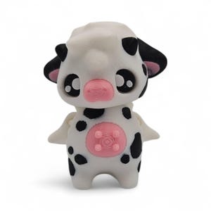 May include: A white and black spotted cow figurine with a pink nose and udder. The figurine has large black eyes and small wings. The cow figurine is a cute and whimsical design.