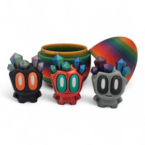 May include: Three colourful, stylised figurines with crystal-like headpieces. The figurines are black, red, and grey, each with large oval eyes in contrasting colours. A rainbow-coloured, curved object and a cylindrical container are in the background.