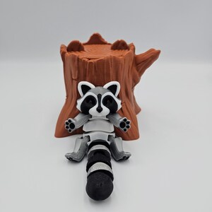 May include: A 3D printed raccoon figurine with a black and white face and a gray and white body. The raccoon is sitting on a brown tree stump.