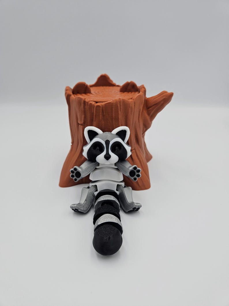 May include: A 3D printed raccoon figurine with a black and white face and a gray and white body. The raccoon is sitting on a brown tree stump.