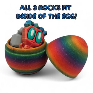 May include: An open, rainbow-hued egg contains a red creature with teal eyes and blue details, plus grey rocks. The text above says "ALL 3 ROCKS FIT INSIDE OF THE EGG!"
