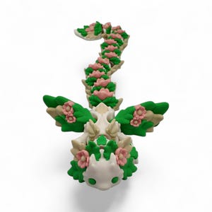 May include: A whimsical, handcrafted sculpture of a dragon, featuring a cream-colored body adorned with green leaves and pink flowers. The dragon has outstretched wings and a curved tail, all decorated with floral accents. The sculpture is set against a white background.