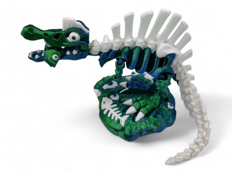 May include: A green and blue dinosaur skeleton toy with white bones. The dinosaur is sitting on a green and blue base with white fish bones.