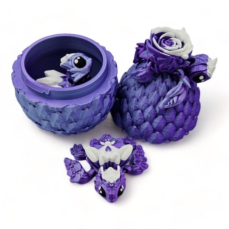 Puede incluir: Un huevo de drag&oacute;n morado se abre para revelar un drag&oacute;n morado m&aacute;s peque&ntilde;o con acentos blancos. El huevo de drag&oacute;n est&aacute; decorado con un dise&ntilde;o texturizado y escamoso. El drag&oacute;n est&aacute; posado con sus alas extendidas.
